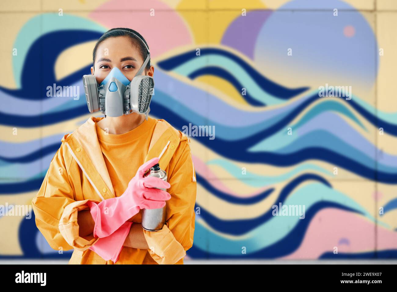 Graffiti female street artist in respirator mask standing near the wall with her paintings looking to camera with copy space. Urban art concept Stock Photo