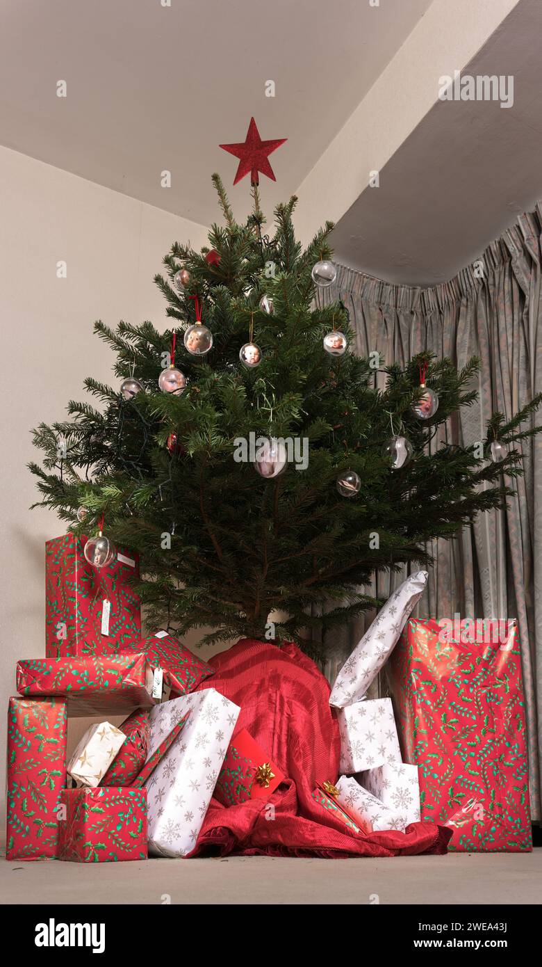 Christmas gifts wrapped in colourful paper stacked disorderly under a christmas tree decorated with baubles and topped with a red star. Stock Photo