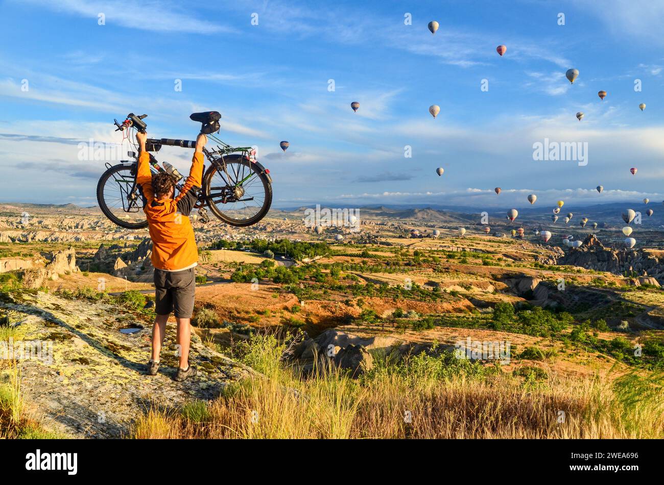 Man lifting his touring bicycle in Cappadocia, in front of the air balloons rising in the early hours of a sunny day Stock Photo