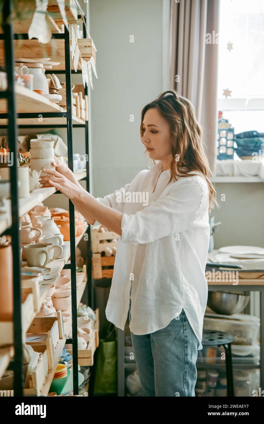 Young woman manager of pottery workshop arranges products on shelf Stock Photo