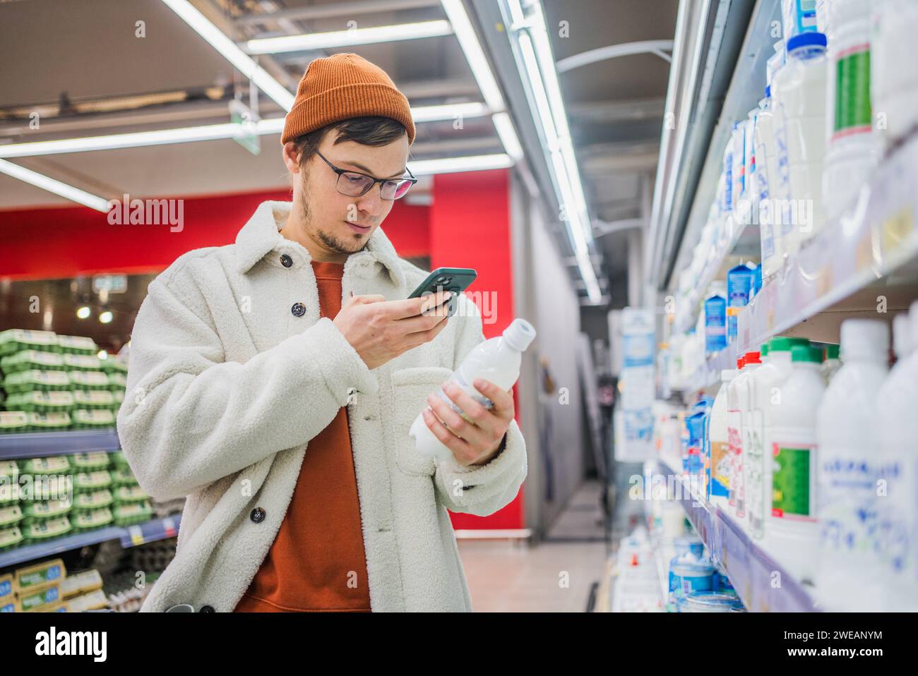 Man in store takes picture of label using phone. Young bearded Caucasian guy in glasses in department with dairy products and eggs holds kefir in hands, takes photo of composition, comparing. Stock Photo