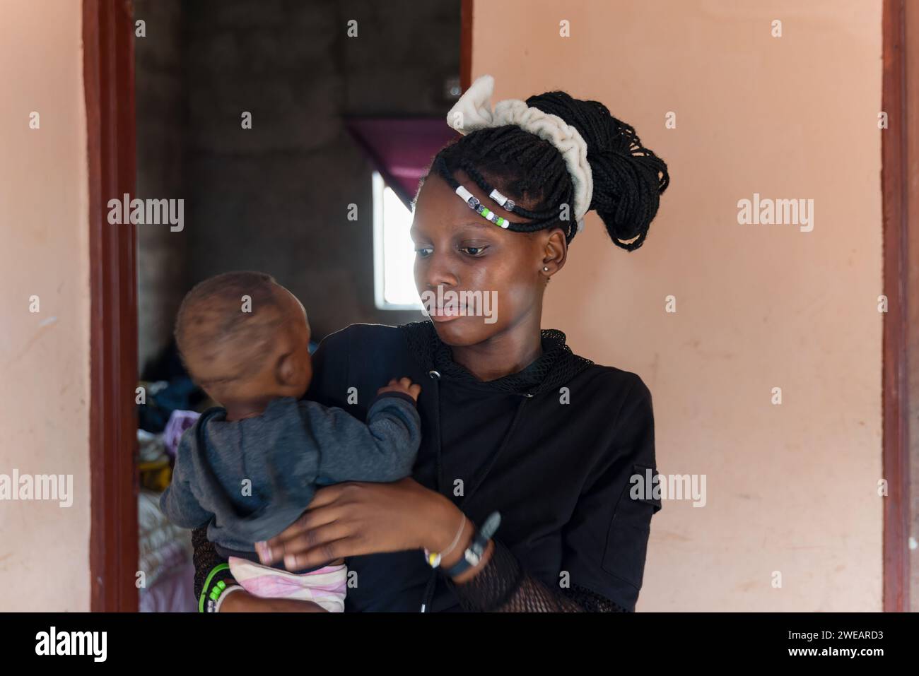 teenage pregnancy, village african girl with braids, holding a baby she is sited next to the door outside, Stock Photo