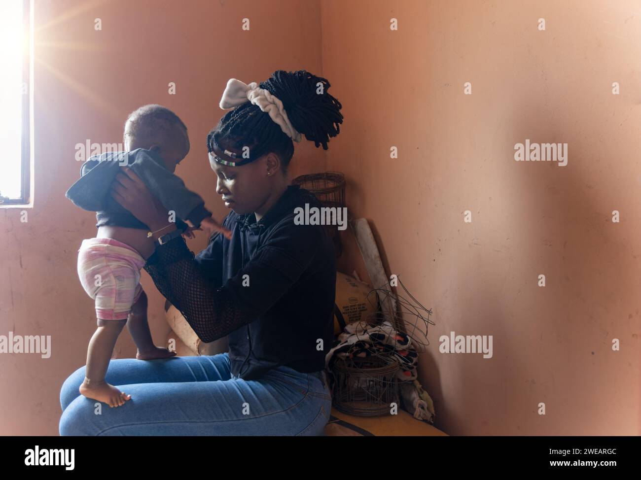 village african girl with braids, holding a baby she is sited next to the window indoors Stock Photo