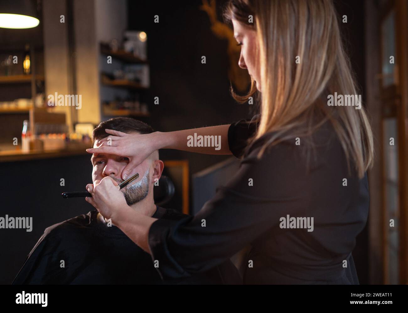 Longhaired young female Barber gently shaving bearded man face bristle