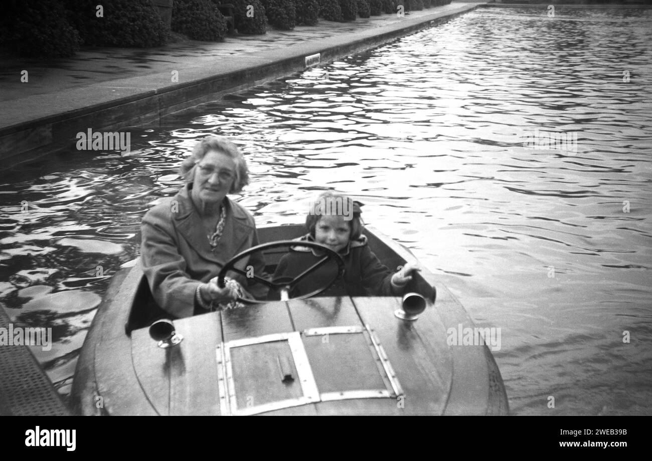 1950s, historical, looking up for a photo, a lady and young girl sitting in small wooden-framed two person motorised boat on a tree-lined boating lake or pond, England, UK. Stock Photo