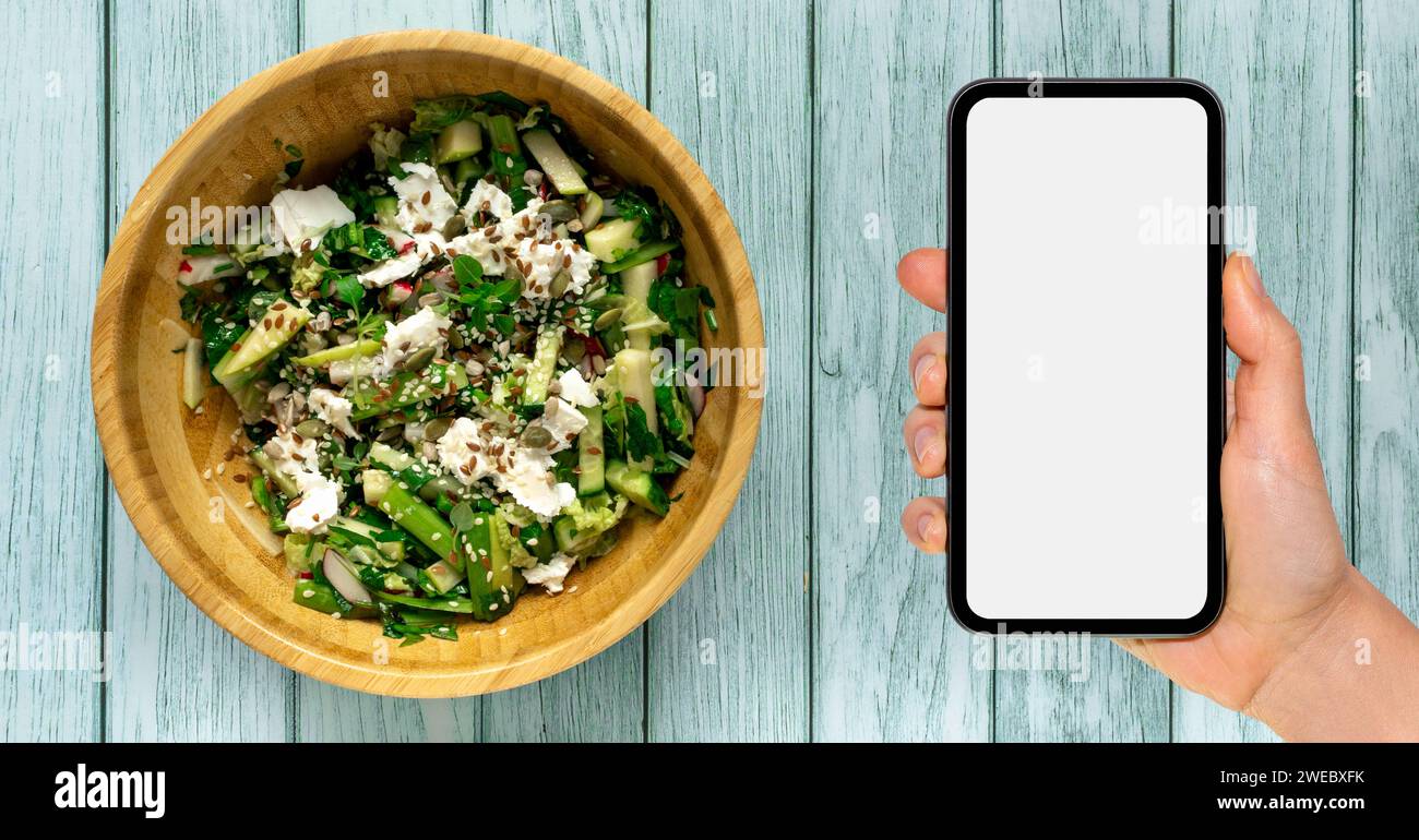 Nutrition mobile app mockup. Smart phone with blank screen in hand next to wooden bowl with green salad. Stock Photo