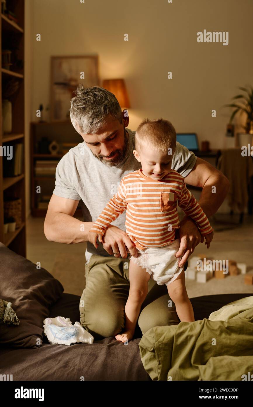 Mature father changing nappy of his adorable baby son standing on bed