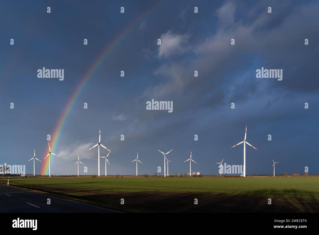Green field with wind farm. Rainbow over the dark sky. Landscape after thunderstorm and rain. Wind turbines illuminated by the sun. Stock Photo