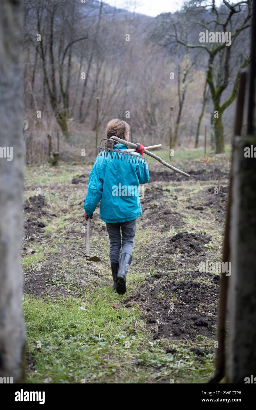 Female Farmer in Blue Cloths and A Rake in Hands Going on Her Garden in Winter to do Some Work Stock Photo