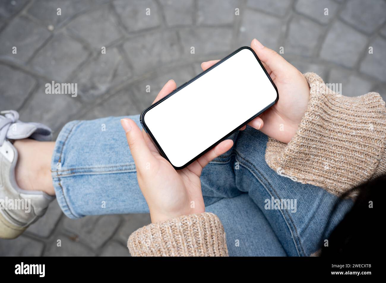 A woman is watching a video on her smartphone while sitting on a bench outdoors, holding a phone in the horizontal position. a white-screen smartphone Stock Photo