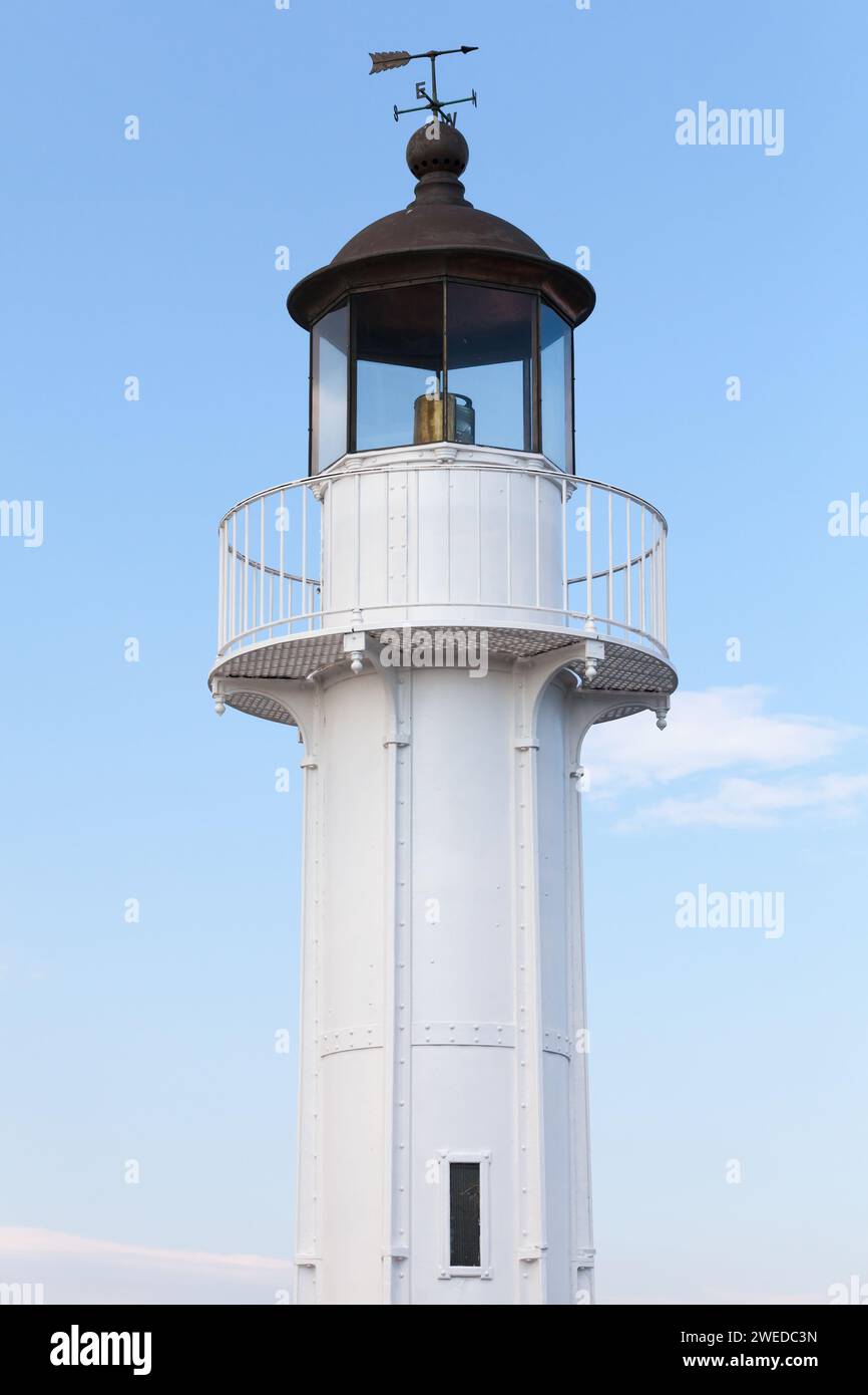 White lighthouse tower with black roof and vane is under blue sky on a daytime, vertical photo. Port of Varna, Bulgaria Stock Photo
