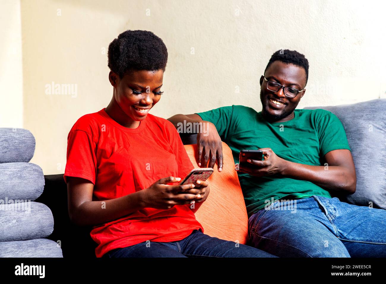 young happy couple sitting on comfortable sofa at home each using mobile phone at home. Stock Photo