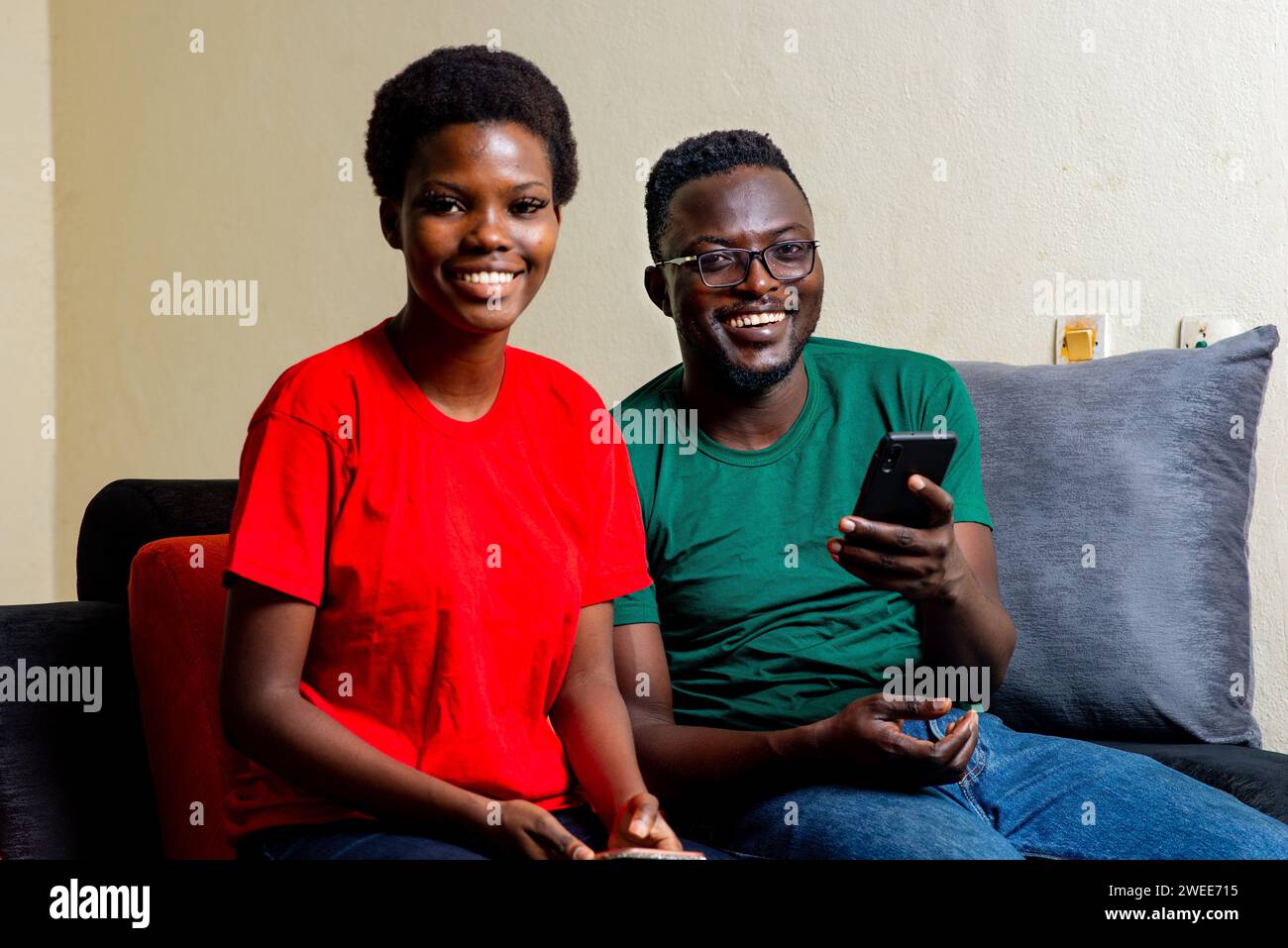 young happy african couple sitting on the comfortable sofa at home on a mobile phone while smiling. Stock Photo