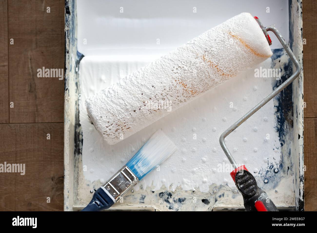 A decorators paint roller and paint brush lying in a paint tray filled with white emulsion paint for covering walls and ceilings with a fresh coat Stock Photo