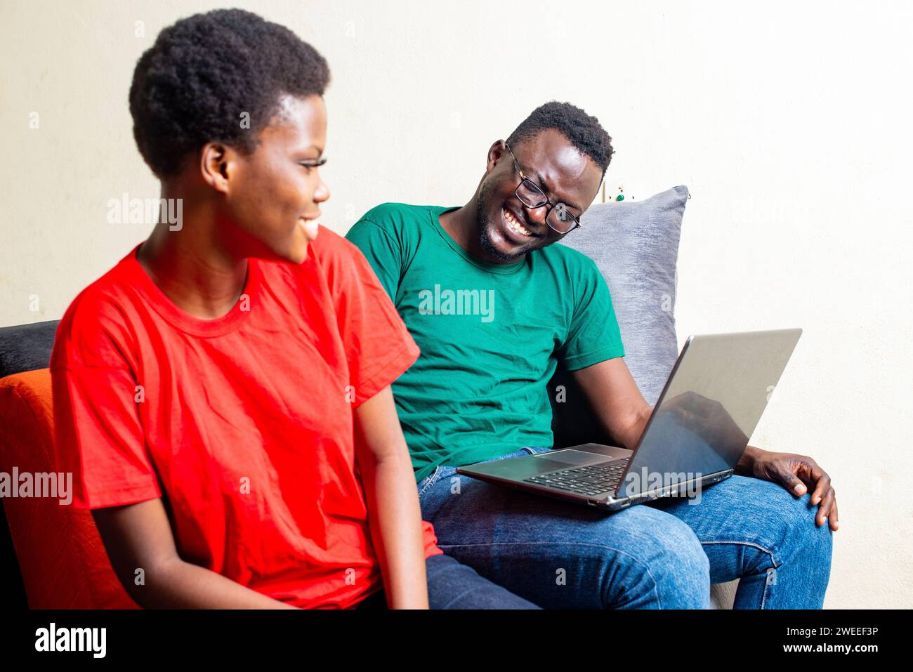 beautiful young happy couple sitting together on sofa at home with laptop while smiling. Stock Photo