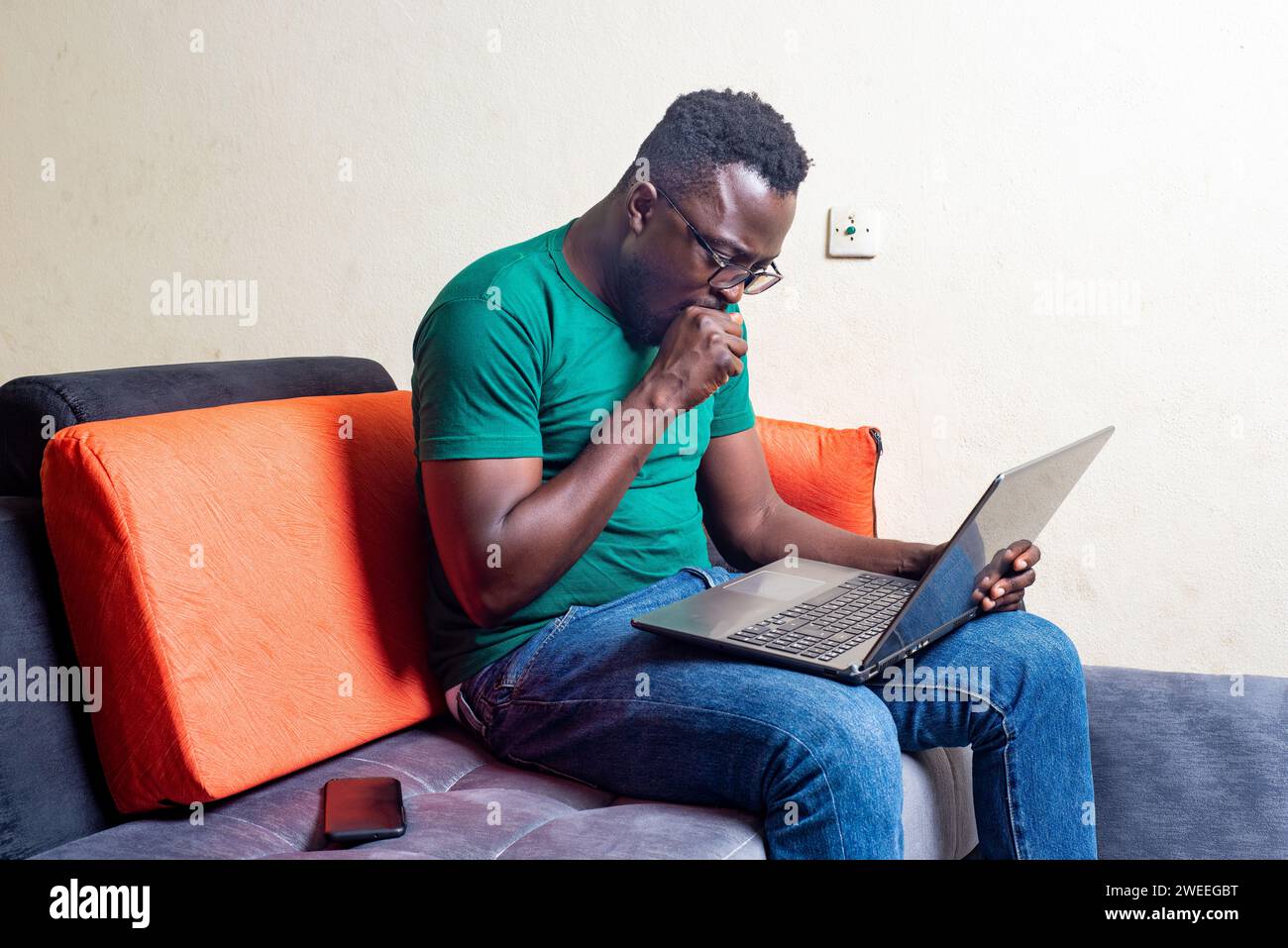 young man in glasses sitting in a sofa home working with laptop covering mouth with fist. Stock Photo