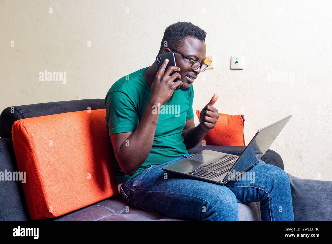 young man in glasses sitting in a sofa home talking on cell phone while using laptop. Stock Photo