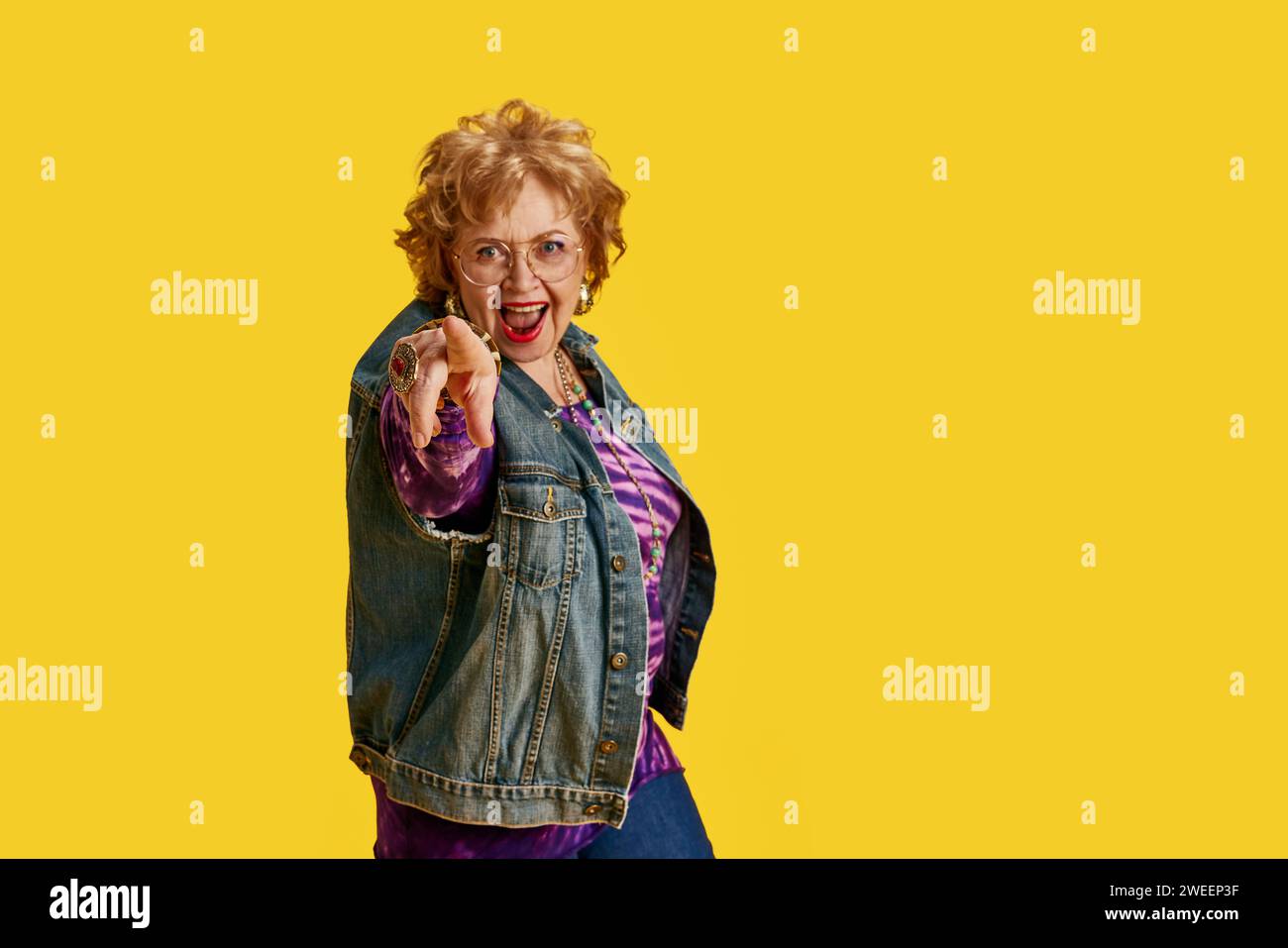Joyful senior woman pointing at camera dressed in denim jacket, purple blouse against yellow background. Stock Photo