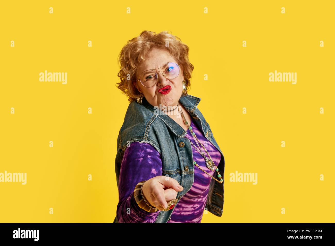 Senior woman puckering lips in round glasses dressed denim jacket, purple T-shirt with gold jewelry against yellow background. Stock Photo