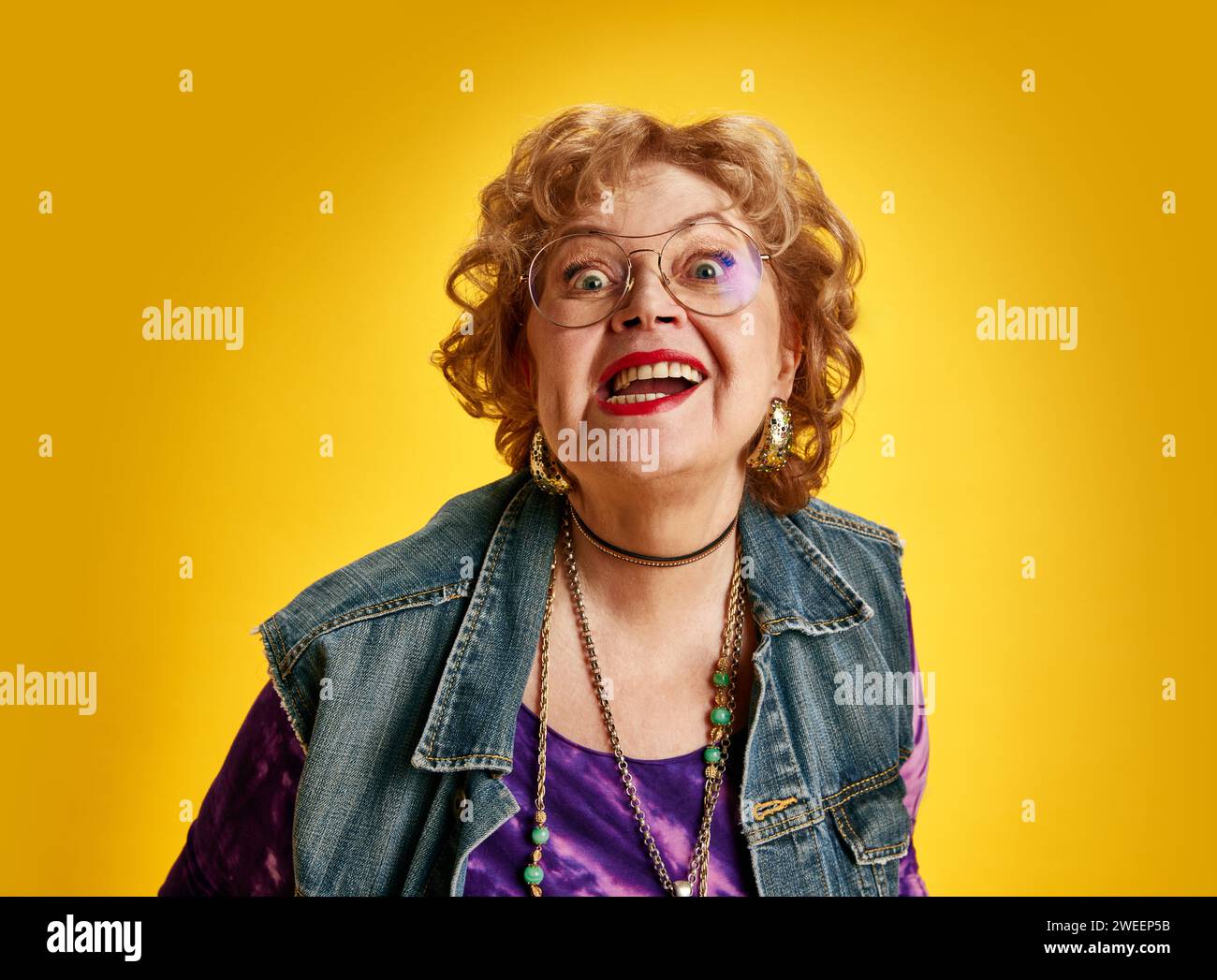 Vibrant smiles at any age. Excited senior lady, wide-eyed in round spectacles dressed denim jacket looking at camera against yellow background. Stock Photo