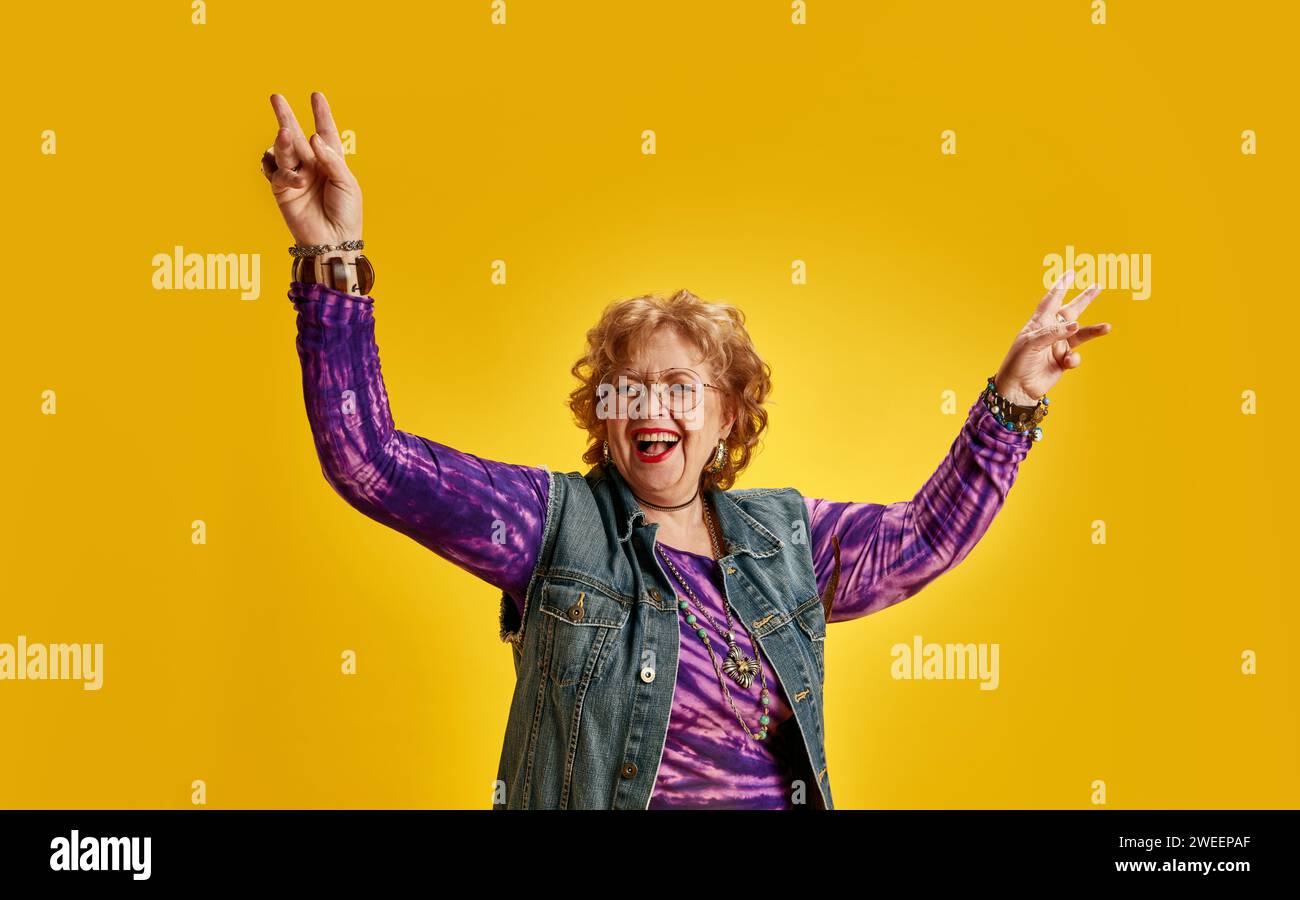 Elderly woman making peace sign and laughing dressed denim jacket and vibrant purple top against yellow background. Happy expression. Stock Photo