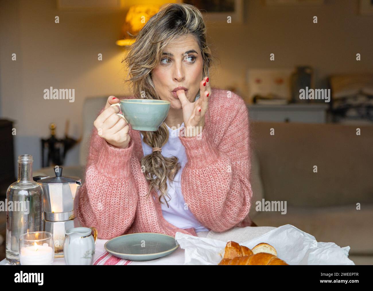 A young woman in a knit sweater appears pensive as she enjoys a cup of coffee, with pastries laid out in front. Stock Photo