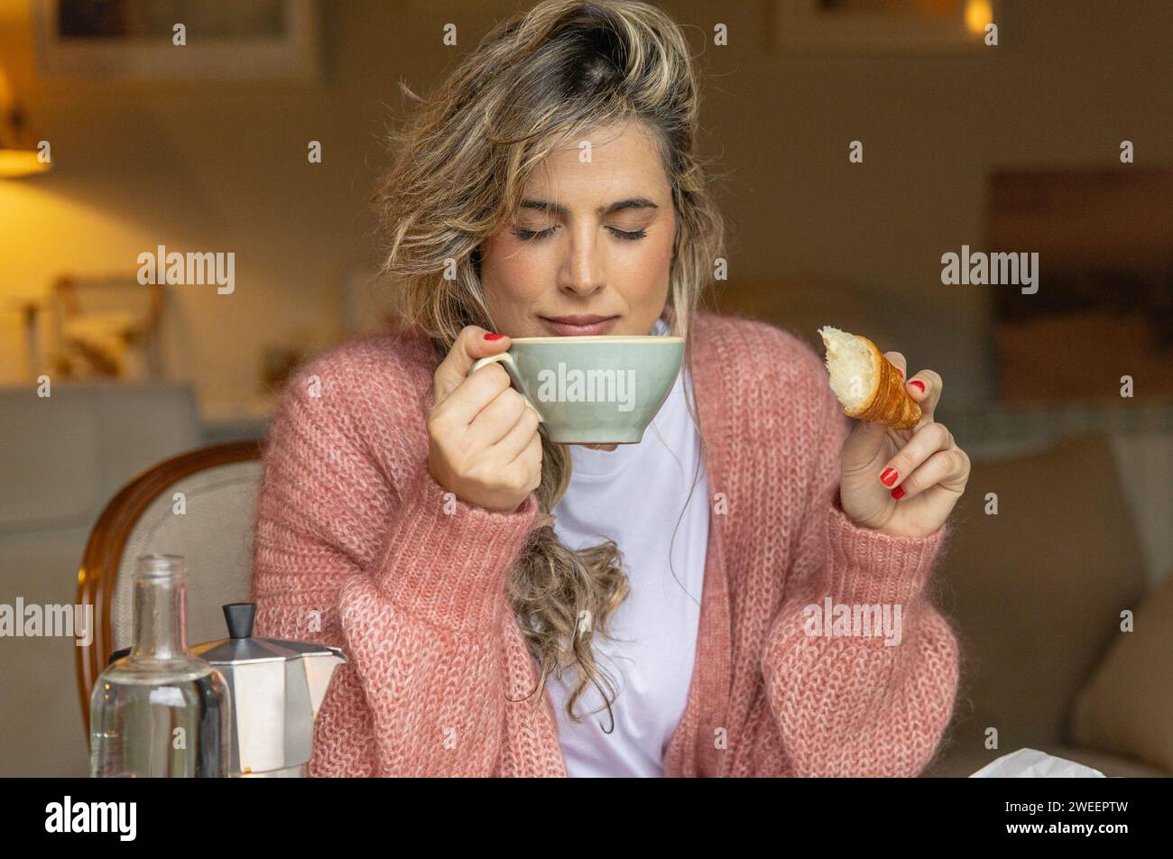A contented young woman in a pink sweater savors the aroma of her coffee with eyes closed, holding a freshly bitten croissant. Stock Photo