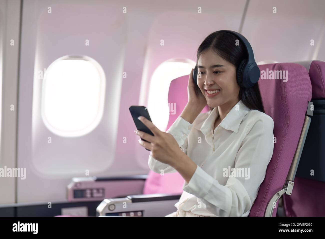 Cheerful Asian woman passenger in headphone watching online movie during intercontinental flight in cabin of aircraft, happy traveler using wifi Stock Photo