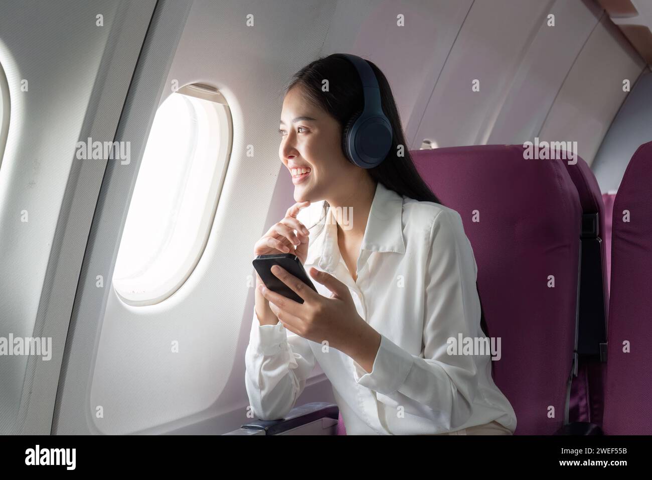 Cheerful Asian woman passenger in headphone watching online movie during intercontinental flight in cabin of aircraft, happy traveler using wifi Stock Photo