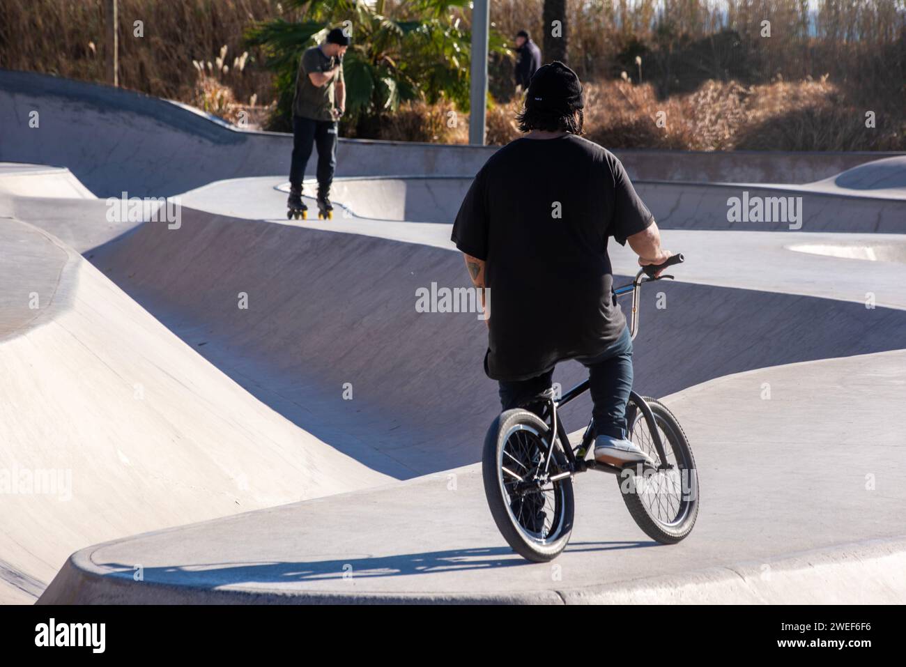 Skate park variety: Biker waits, skater glides in a diverse urban scene Stock Photo