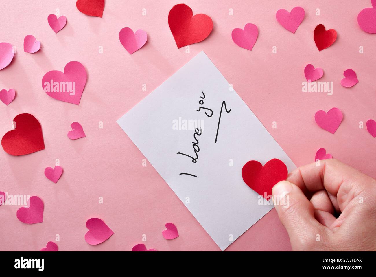Romantic note with writing I love you on white paper on pink background with hand with a red heart cutout and many hearts around. Top view. Stock Photo