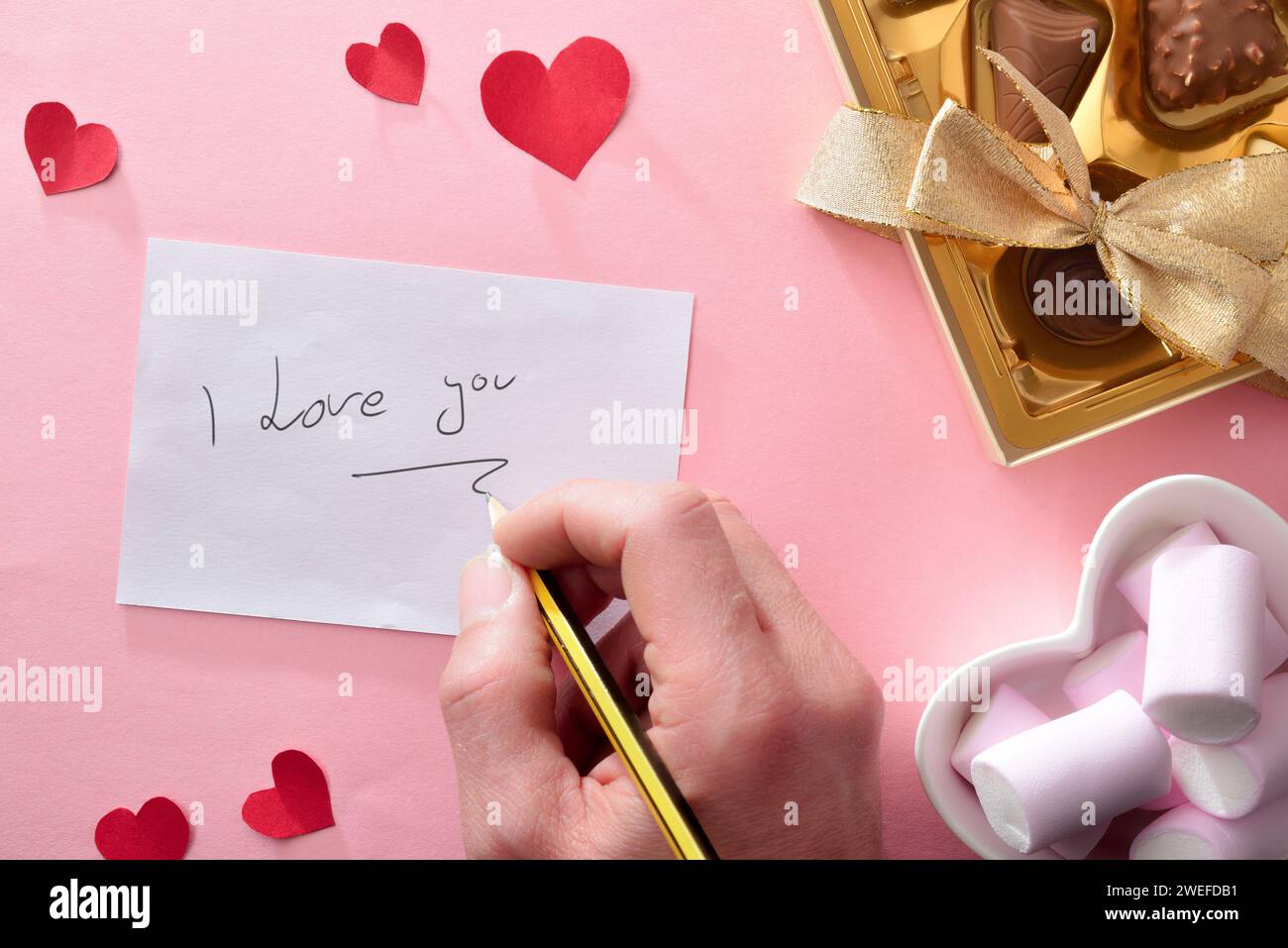 Hand writing I love you on white sheet on pink background with box of chocolates and container with sweets. Top view. Stock Photo