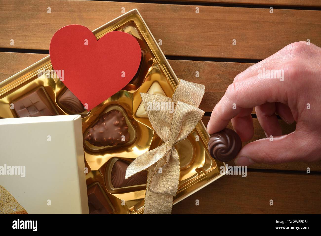 Gift box with golden bow full of assortment of chocolates on wooden table and hand taking a chocolate to try. Top view. Stock Photo