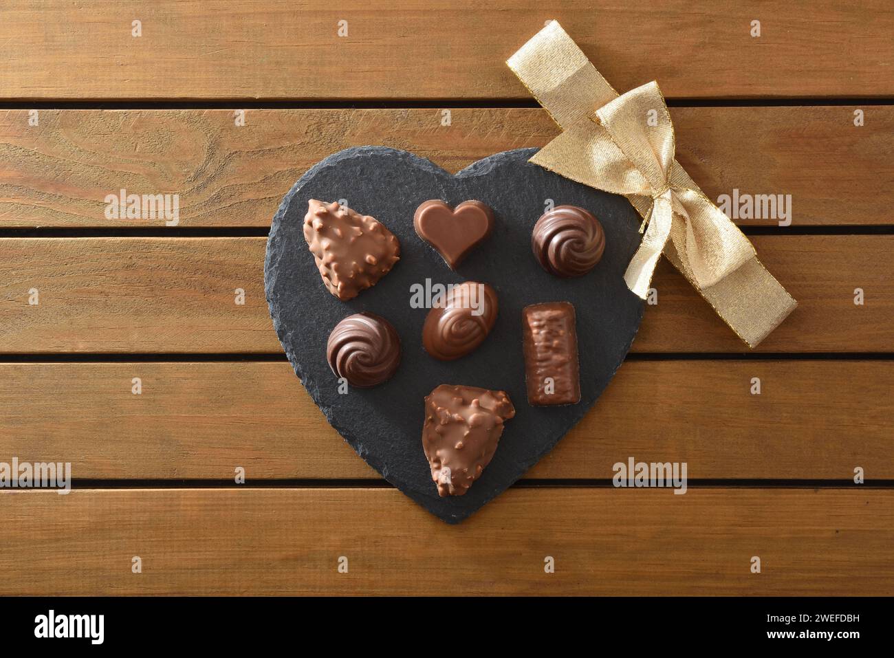 Assortment of chocolates on heart-shaped slate plate with golden bow in the center on wooden slatted table. Top view. Stock Photo