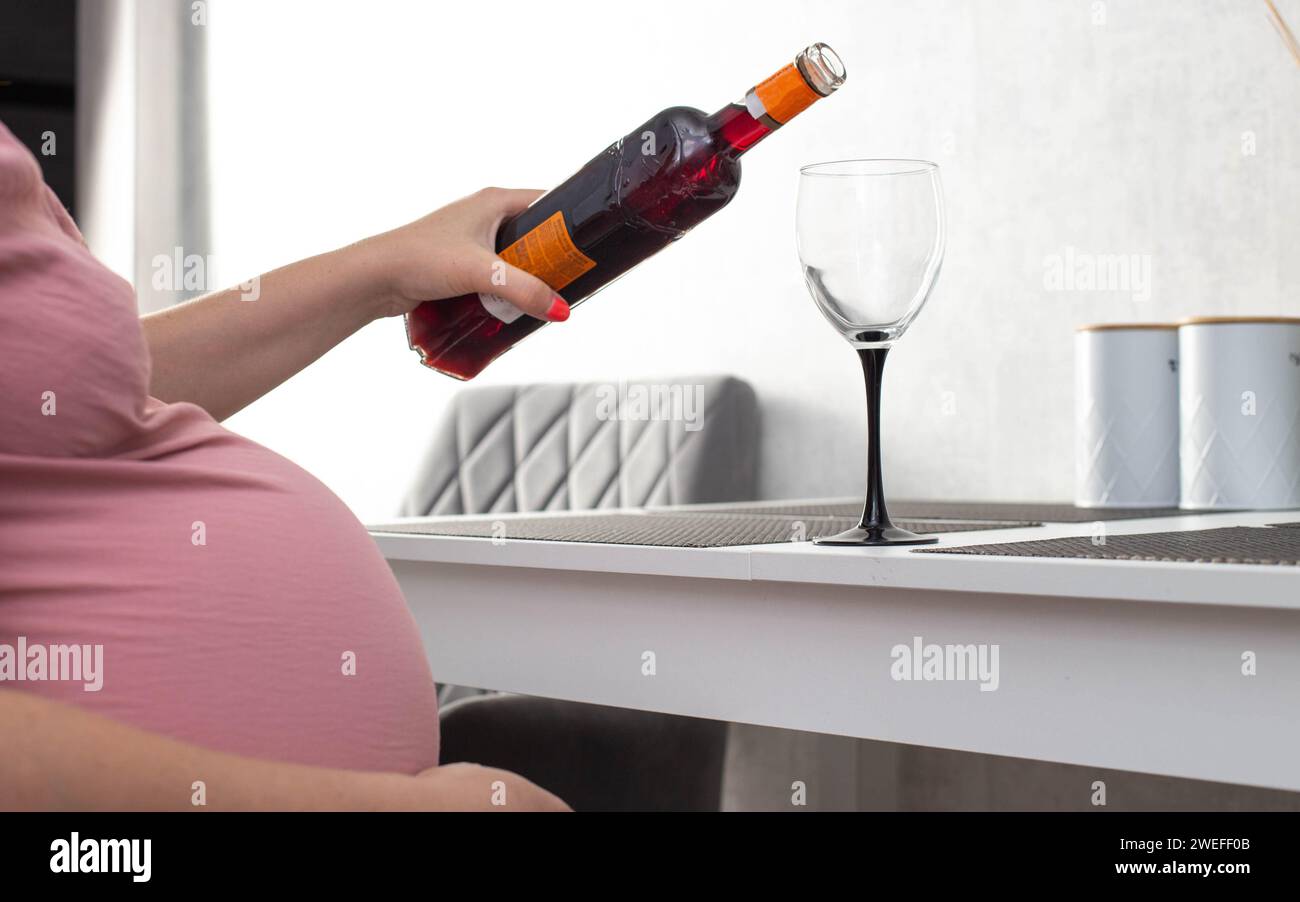 A pregnant girl pours wine from a bottle into a glass. The concept of drinking alcohol during pregnancy. Harm and cognitive impairment in a child. Alc Stock Photo
