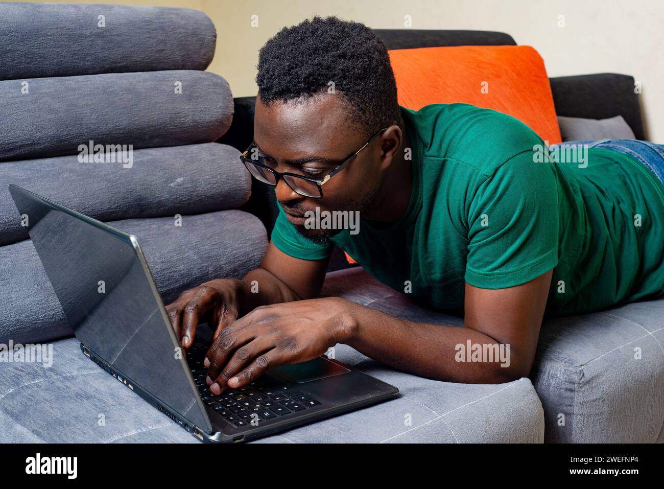 young man wearing his optical glasses and lying on a sofa at home while working with the laptop. Stock Photo