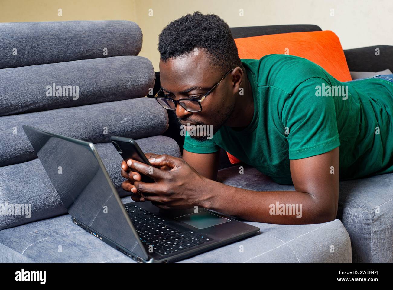 young man wearing optical glasses lying on sofa at home uses mobile phone and laptop. Stock Photo