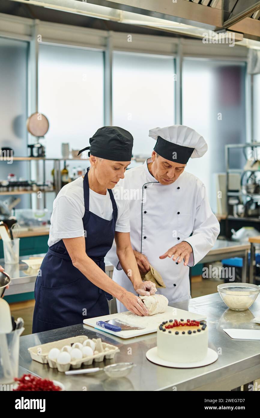 attractive mature chef in apron and toque working with dough next to her chief cook, confectionary Stock Photo