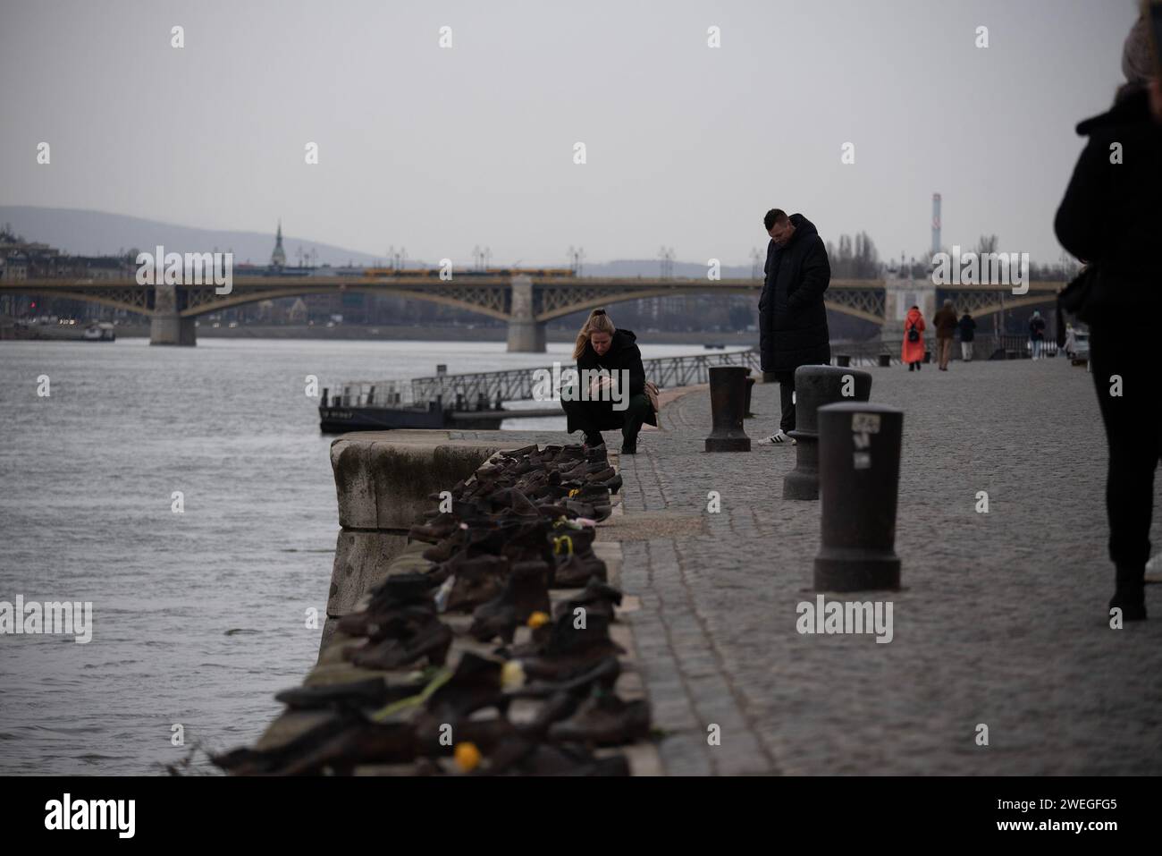 Budapest, Hungary. 24th Jan, 2024. A woman takes photos of the shoes on the bank of Danube River and Margaret Bridge in the background. A Sculpture of Shoes on the bank of river Danube was formed in 2005 on the East bank of the river by the Gyula Pauer. To honor the Jewish people who were shot into the river by the fascist Hungarian militia called Arrow Cross Party in 1944-45. It became a main attraction for the tourists and the people who want to show respect to the victims. (Photo by Krisztian Elek/SOPA Images/Sipa USA) Credit: Sipa USA/Alamy Live News Stock Photo