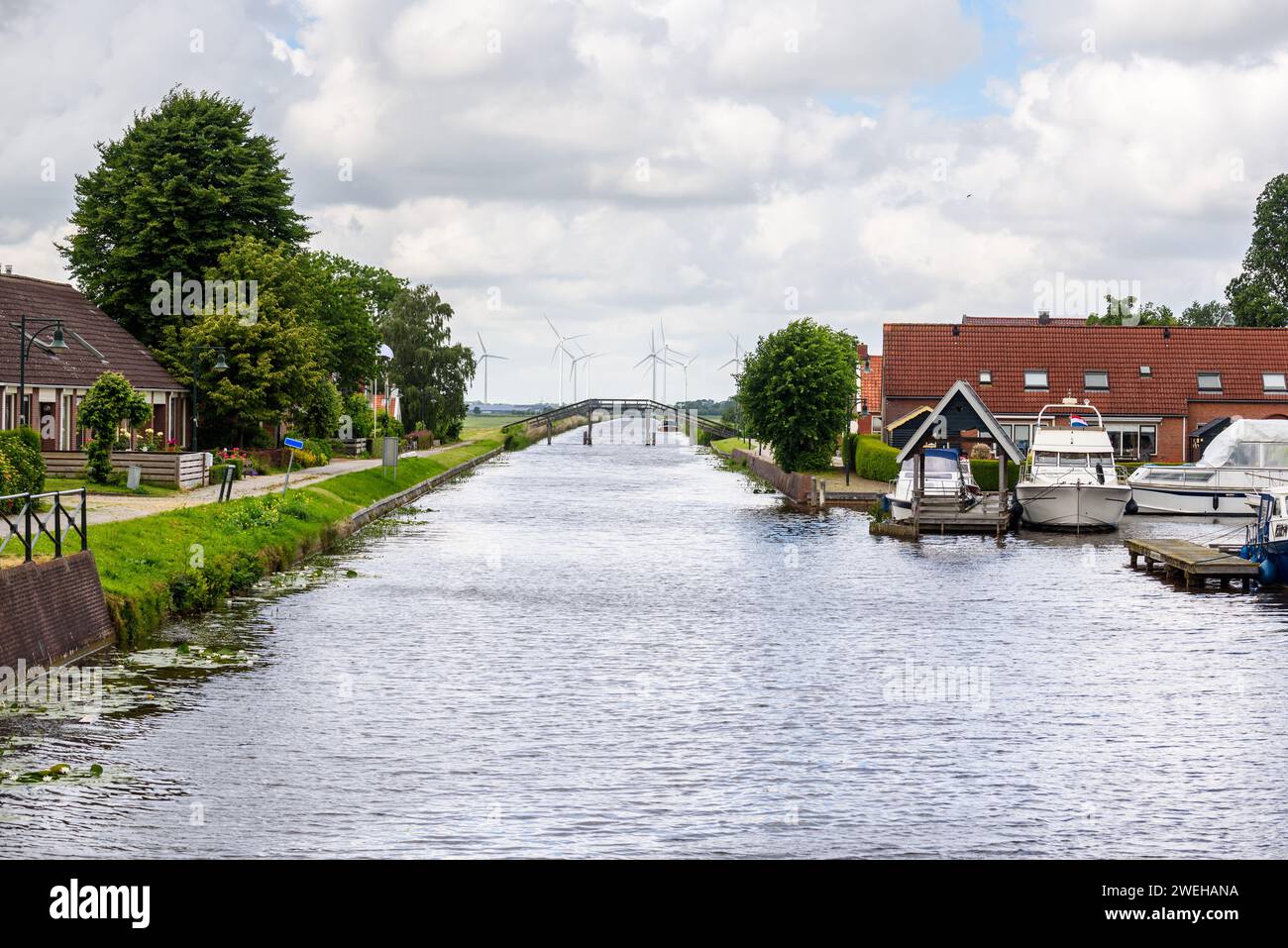 Canal through a small town in the countryside of Netherland on an overcast summer day. A wind farm is visible in background. Stock Photo
