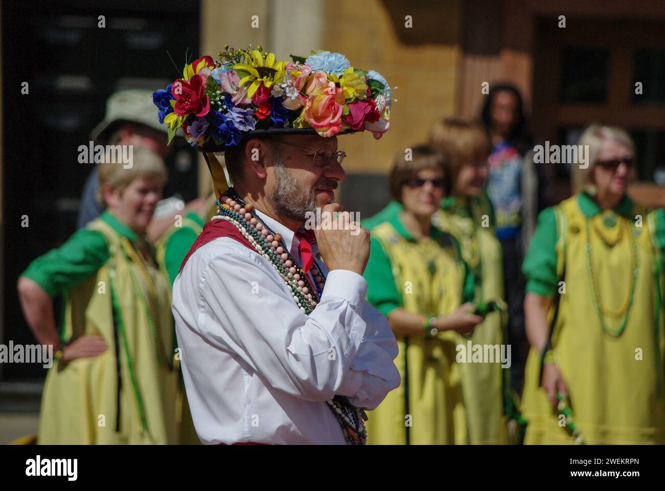 Morris Man wearing a hat decorated with flowers at a Morris Dancing convention, Northampton, UK; out of focus lady Morris Dancers to the rear Stock Photo