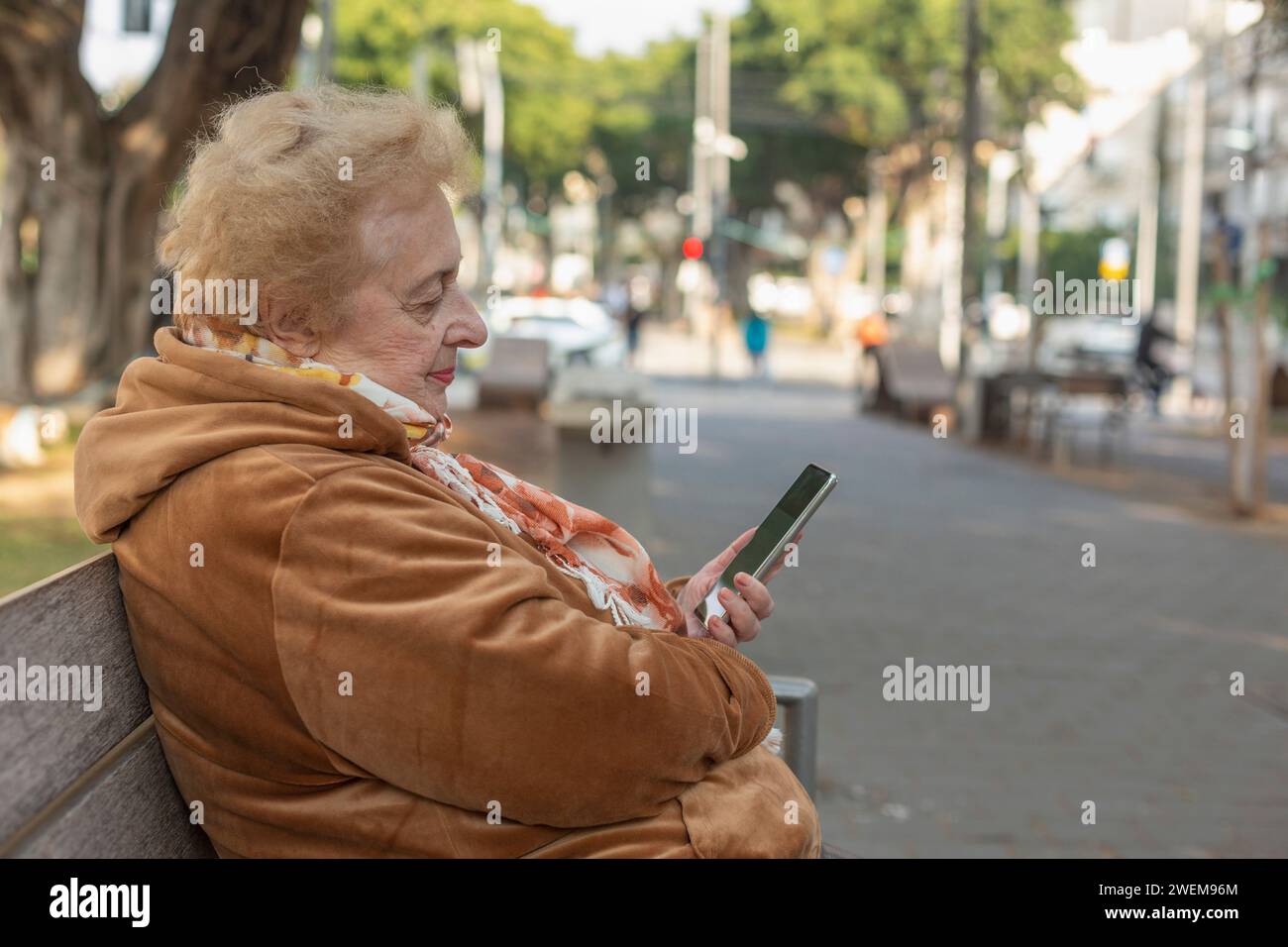 Senior woman sitting and using smart phone on city sidewalk bench Stock Photo