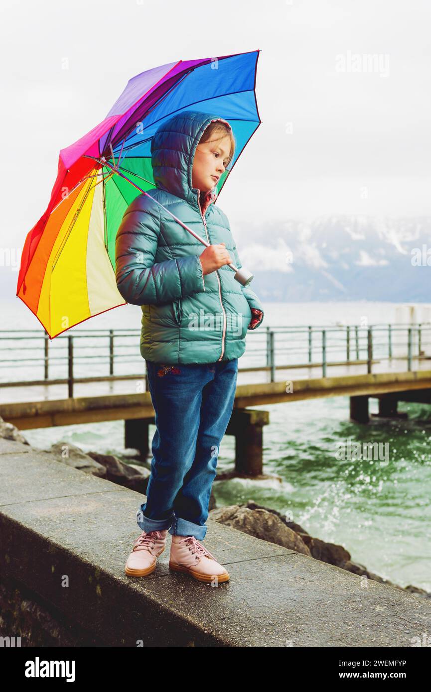 Outdoor portrait of a cute little girl, wearing warm green jacket, holding big colorful umbrella, standing next to lake Geneva, Switzerland Stock Photo