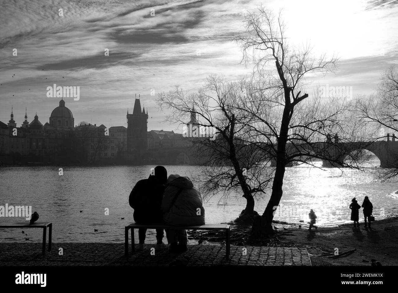 Loving couple on a bench, looking across the river in Prague as a family play on the river bank and sunlight hits the water Stock Photo