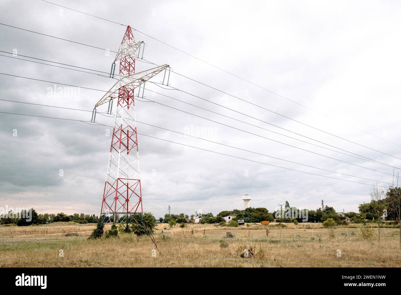 A towering electricity pylon stands against a cloudy sky, with multiple high-voltage power lines extending into the distance over a rural landscape. Stock Photo