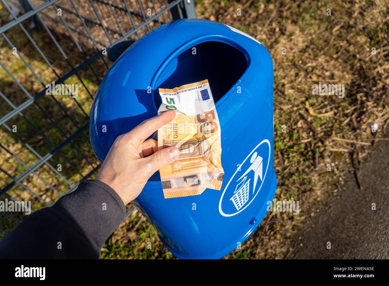 Langweid, Bavaria, Germany - January 26, 2024: Man throws euro cash banknotes into a blue trash can. Symbolic image of loss of value of money, recession and inflation *** Mann wirft Euro Bargeld Geldscheine in einen blauen Mülleimer. Symbolbild Wertverlust von Geld, Rezession und Inflation Stock Photo