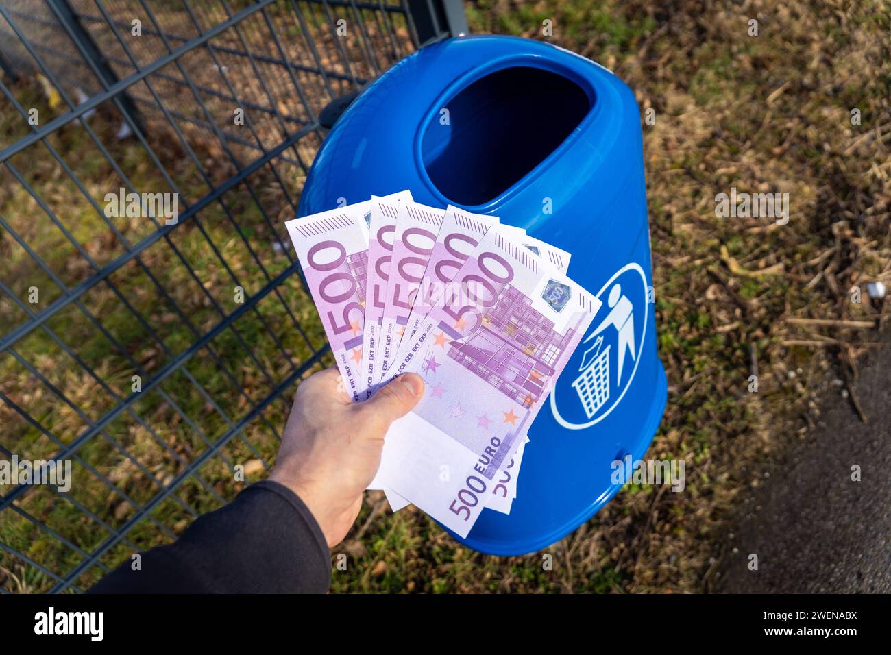 Langweid, Bavaria, Germany - January 26, 2024: Man throws euro cash banknotes into a blue trash can. Symbolic image of loss of value of money, recession and inflation *** Mann wirft Euro Bargeld Geldscheine in einen blauen Mülleimer. Symbolbild Wertverlust von Geld, Rezession und Inflation Stock Photo