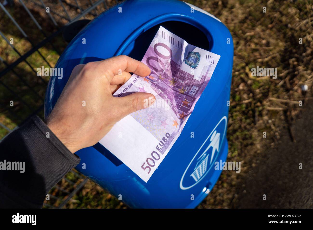 Langweid, Bavaria, Germany - January 26, 2024: Man throws euro cash banknotes into a blue trash can. Symbolic image of loss of value of money, recession and inflation *** Mann wirft Euro Bargeld Geldscheine in einen blauen Mülleimer. Symbolbild Wertverlust von Geld, Rezession und Inflation Stock Photo