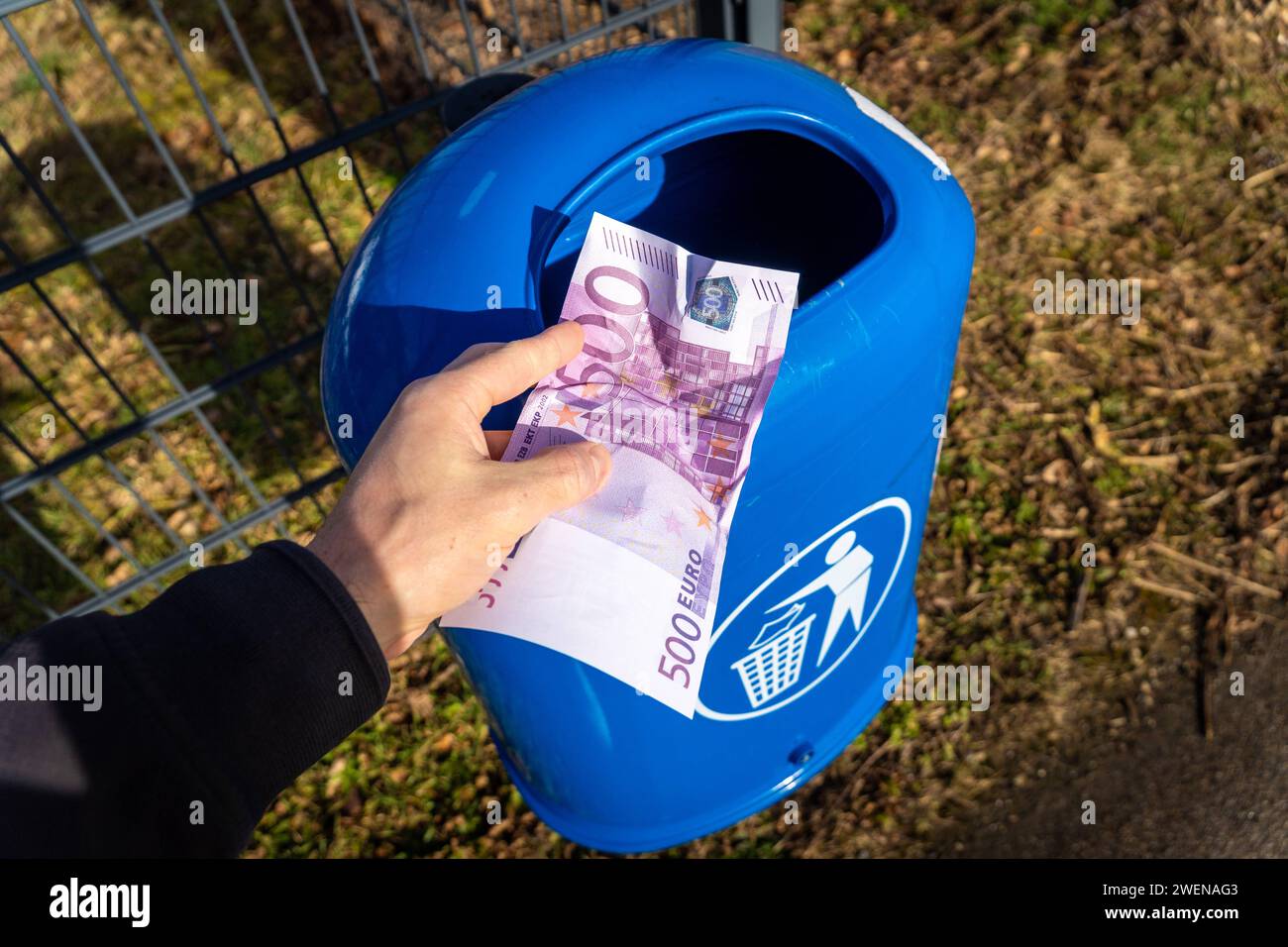 Langweid, Bavaria, Germany - January 26, 2024: Man throws euro cash banknotes into a blue trash can. Symbolic image of loss of value of money, recession and inflation *** Mann wirft Euro Bargeld Geldscheine in einen blauen Mülleimer. Symbolbild Wertverlust von Geld, Rezession und Inflation Stock Photo