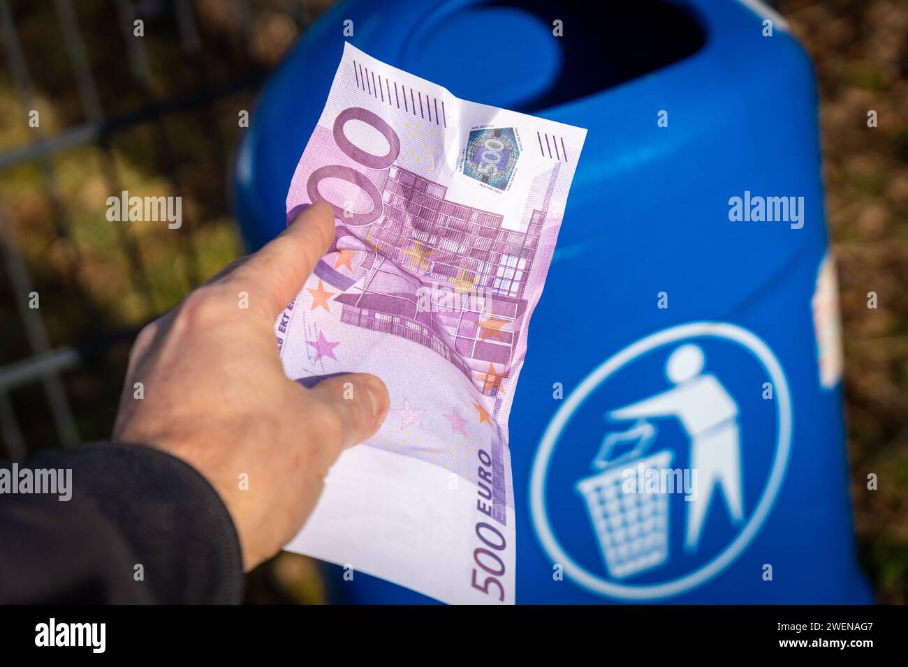 Langweid, Bavaria, Germany - January 26, 2024: Man throws euro cash banknotes into a blue trash can. Symbolic image of loss of value of money, recession and inflation *** Mann wirft Euro Bargeld Geldscheine in einen blauen Mülleimer. Symbolbild Wertverlust von Geld, Rezession und Inflation Stock Photo